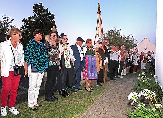 Eine Gruppe von Menschen steht draußen in der Nähe einer Kirche. Einige sind in traditioneller Kleidung gekleidet. Es gibt Blumen und einen Pfad in der Nähe. Bäume und ein Gebäude sind im Hintergrund zu sehen.
