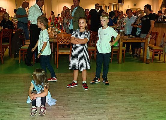 Eine Zusammenkunft von Erwachsenen und Kindern in einem Raum mit Holzboden. Drei Kinder stehen vorne, während ein Mädchen auf dem Boden sitzt. Der Raum hat Holztische und Stühle.