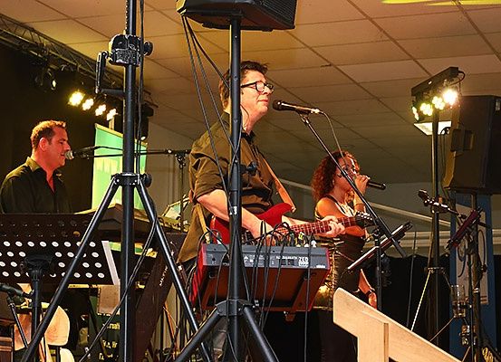 Eine Band spielt auf der Bühne mit Musikinstrumenten unter Bühnenbeleuchtung, ein Mann spielt Gitarre und singt in ein Mikrofon.