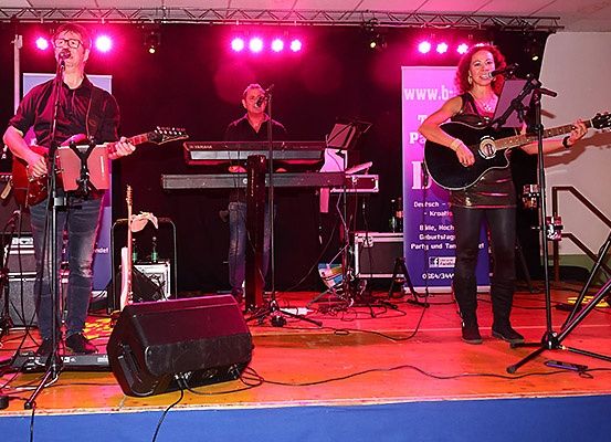 Eine Band tritt mit roter Beleuchtung auf der Bühne auf. Ein Mann spielt Gitarre und singt, ein anderer spielt Keyboard und eine Frau spielt Gitarre und singt. Sie sind von Ausrüstung umgeben.