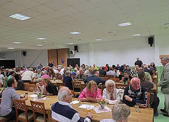 Eine große Gruppe von Menschen sitzt an langen Tischen in einem Saal. Sie scheinen in Gespräche vertieft zu sein. Der Raum ist gut beleuchtet, mit Tischen, die mit Gläsern, Blumen und Papieren bedeckt sind. Einige Personen stehen in der Nähe der Tische. Der Saal hat ein modernes Design mit einer weißen Decke und Wänden.