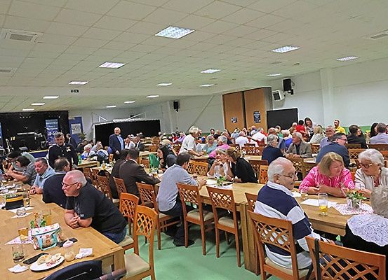 Ein großer Saal mit Menschen an langen Tischen, einige mit Brillen und andere in Gesprächen vertieft. Auf den Tischen stehen Getränke, Essen und Blumen. Eine Bühne mit Lautsprecher im Hintergrund.