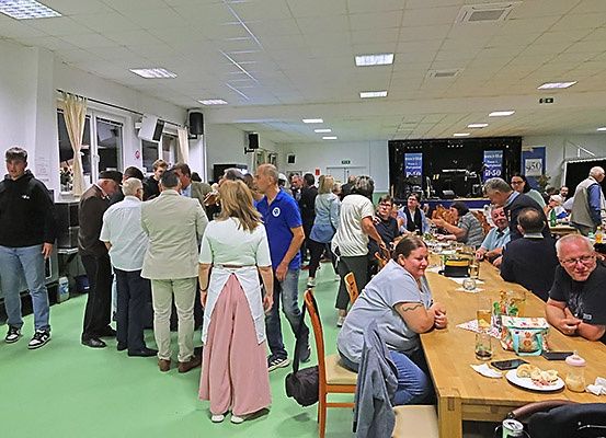 Ein Raum voller Menschen beim Essen und Plaudern; einige stehen und andere sitzen um lange Tische mit Essen und Getränken.