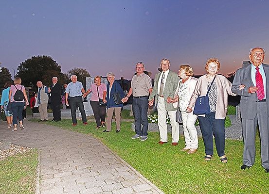 Eine Gruppe formell gekleideter Menschen, die Händchen halten, steht auf einem Rasenfeld. Dahinter befindet sich ein Weg, ein Gebäude und ein Baum. Der Himmel ist blau mit einigen Wolken.