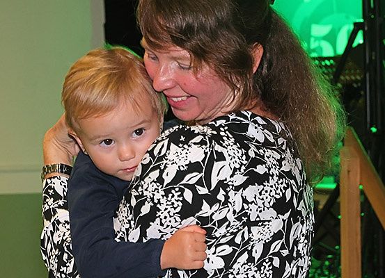 Eine Frau mit langen Haaren lächelt und umarmt einen jungen Jungen mit blonden Haaren. Sie stehen eng beieinander in einem Raum.