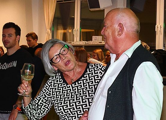 Eine ältere Frau mit Brille hält ein Weinglas und unterhält sich mit einem älteren Mann bei einer sozialen Veranstaltung. Hinter ihnen unterhalten sich andere Menschen.
