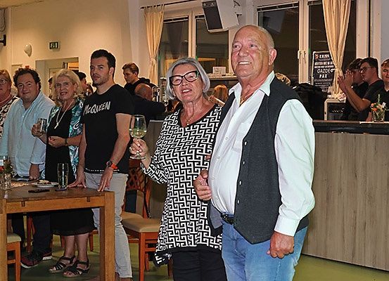 Eine Gruppe von Menschen versammelt sich drinnen, einige halten Weingläser. Eine Frau und ein Mann stehen in der Mitte und lächeln. Hinter ihnen ist eine Theke zu sehen, und weitere Menschen stehen und klatschen.