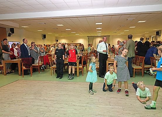 Eine Gruppe von Menschen steht und sitzt in einem Saal. Einige Leute stehen vorne, und einige sitzen auf Stühlen. Eine Person steht in der Mitte und scheint zu sprechen. Ein Tisch ist hinter ihnen aufgestellt. Einige Lautsprecher sind an der Wand montiert. Ein Monitor ist auf dem Schreibtisch platziert.