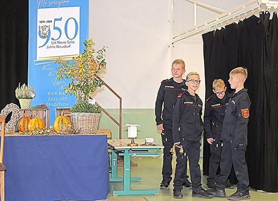 Fünf Jungen in Uniform stehen vor einer Bühne, einer hält eine Glocke. Dahinter steht ein Tisch mit Kürbissen und einem Schild, das 'Hier hat Zukunft Tradition' besagt. Der Hintergrund zeigt '950 Jahre Neudorf'.