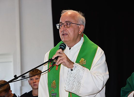 Ein älterer Priester in einer grünen Stola spricht in ein Mikrofon, möglicherweise während einer religiösen Zeremonie. Hinter ihm stehen einige Kinder und ein weiterer Priester.
