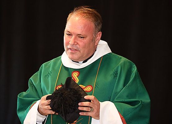 Ein Priester hält einen schwarzen Hut mit einem Bommel in den Händen, gekleidet in ein grünes Gewand.