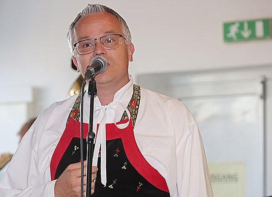 Ein Mann mit Brille und einer traditionellen Schürze singt in ein Mikrofon in einem Raum mit weißen Wänden und einem Ausgangsschild.