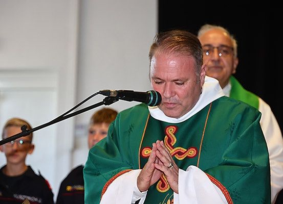 Ein Priester betet mit zusammengepressten Händen vor einem Mikrofon. Hinter ihm sind zwei weitere Männer anwesend, einer von ihnen trägt eine Brille.