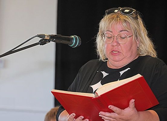 Eine Frau mit Brille und schwarzem Shirt liest ein rotes Buch vor einem Mikrofon. Sie hat blonde Haare und steht vor einem schwarzen Hintergrund.