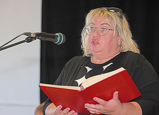 Eine Frau mit Brille und schwarzem Shirt steht vor einem Mikrofon und liest aus einem roten Buch. Hinter ihr ist ein schwarzer Hintergrund zu sehen.