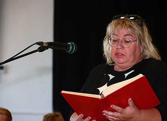 Eine Frau mit Brille liest ein rotes Buch vor einem Mikrofon. Sie trägt ein schwarzes Shirt. Hinter ihr befindet sich eine weiße Wand.