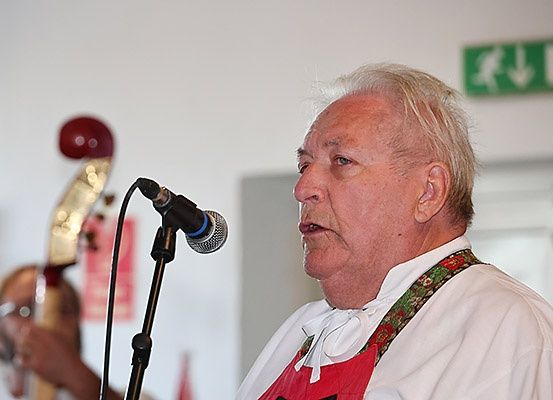 Ein älterer Mann singt in ein Mikrofon und trägt eine rote Schürze. Hinter ihm spielt eine andere Person ein Saiteninstrument. Ein Notausgangsschild ist an der Wand zu sehen.