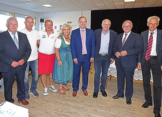 Eine Gruppe von Personen, die formell gekleidet sind, darunter Männer in Anzügen und Krawatten und eine Frau in einem traditionellen Kleid, stehen in einem Raum.