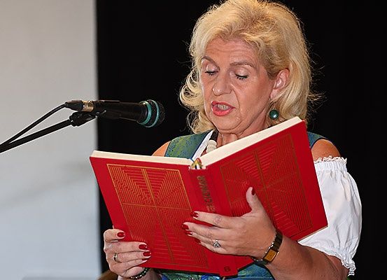 Eine Frau mit blondem Haar, die ein grünes und weißes Kleid trägt, liest ein rotes Buch vor einem Mikrofon.