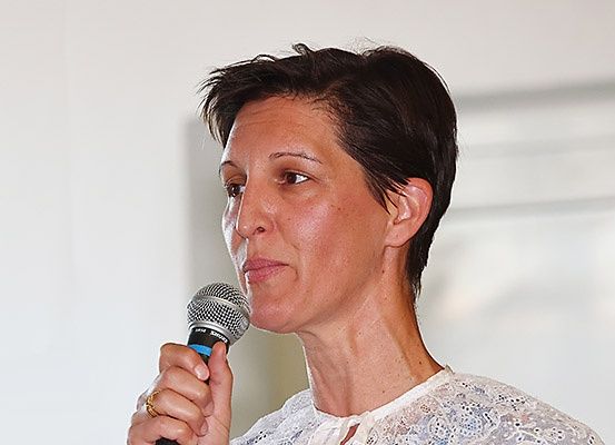 Eine Frau mit kurzen Haaren und einem weißen Spitzenoberteil spricht in ein Mikrofon, möglicherweise bei einer Veranstaltung.