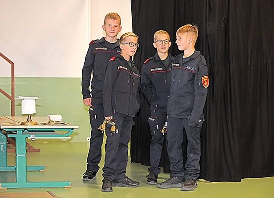Vier Jungen stehen in einem Raum in gleichen Uniformen, einer hält eine Glocke. Hinter ihnen steht ein Tisch mit einer Lampe.
