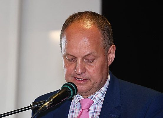 Ein Mann in einem blauen Anzug und einer rosa Krawatte spricht in ein Mikrofon. Er hat einen ernsten Gesichtsausdruck und seine Augen sind geschlossen.