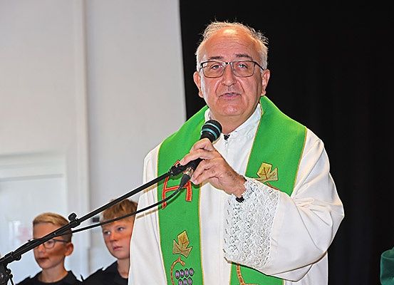 Ein Priester steht an einem Mikrofon und spricht zu einem Publikum. Er trägt eine grün-weiße Stola. Hinter ihm schauen zwei Jungen zu. Die Umgebung scheint eine Kirche zu sein.