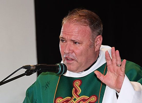 Ein Priester hält eine Rede bei einer Zeremonie, trägt ein grünes Gewand mit Kreuzdesign und einen Ring. Er hebt die Hand, möglicherweise eine Geste machend.