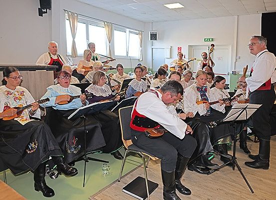 Eine Gruppe von Musikern, einige in traditioneller Kleidung, sitzt und spielt verschiedene Saiteninstrumente. Ein Mann steht in ihrer Nähe, möglicherweise dirigiert er. Der Innenraum ist mit Holzboden und weißen Wänden ausgestattet.