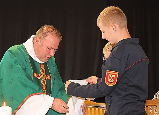 Ein Priester reicht einem jungen Jungen ein weißes Tuch, möglicherweise während einer religiösen Zeremonie, mit einer Kerze und einem schwarzen Hintergrund hinter ihnen.