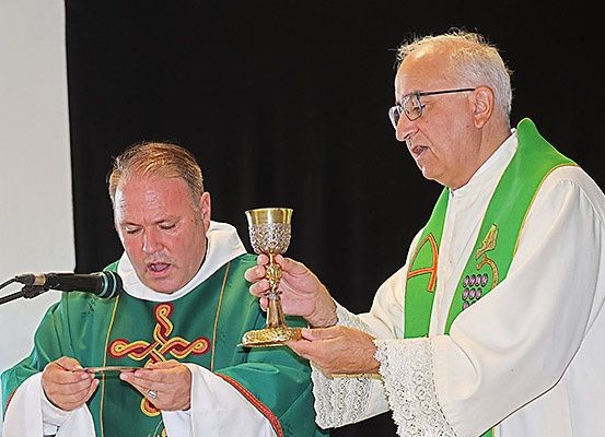 Zwei Priester in grünen Gewändern halten einen Kelch und eine Hostie, wahrscheinlich während einer religiösen Zeremonie.
