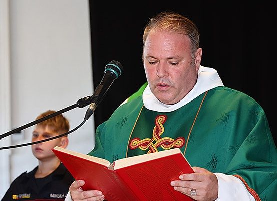 Ein Priester liest aus einem Buch, während er einen grünen Talar trägt und ein Mikrofon vor einer weißen Wand und einem Jungen in einer schwarzen Uniform hält.