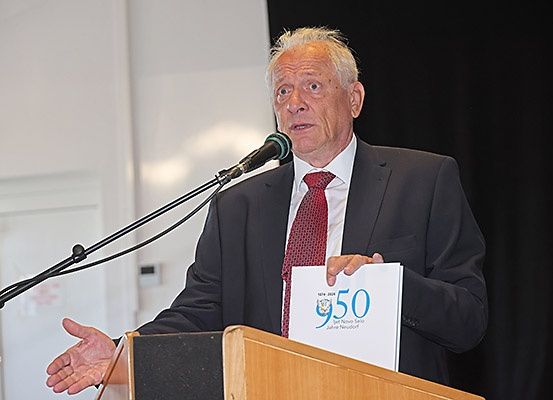 Ein älterer Mann in Anzug und Krawatte spricht an einem Rednerpult und hält ein Papier mit der Nummer 950 und den Worten 'New Now' in der Hand.