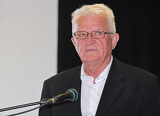 Ein älterer Mann mit Brille und einem schwarzen Anzug steht an einem Rednerpult und sieht ernst aus, während er in ein Mikrofon spricht.