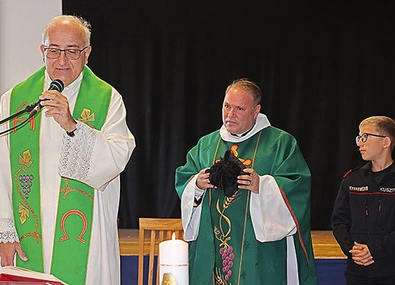 Zwei Priester stehen vor einem Mikrofon, einer hält ein Buch, der andere einen Kelch, mit einem jungen Jungen in Uniform und einer Kerze auf einem Tisch im Hintergrund.