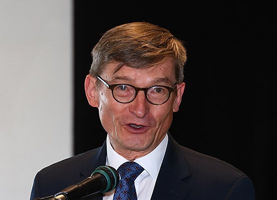 Ein Mann in formeller Kleidung spricht in ein Mikrofon. Er trägt eine Brille und eine Krawatte und steht vor einem schlichten Hintergrund.