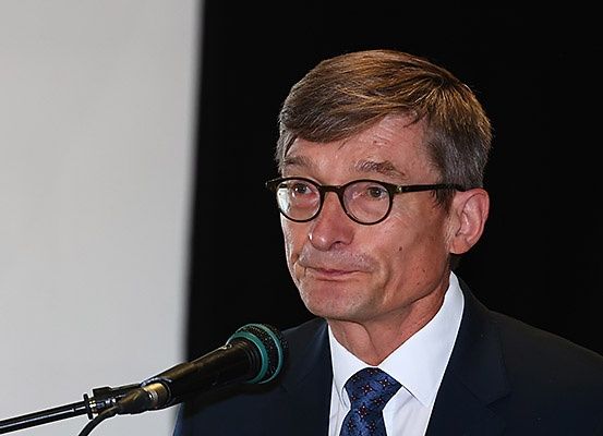 Ein Mann in Anzug und Krawatte steht hinter einem Mikrofon, möglicherweise bei einer Rede. Er trägt eine Brille und hat einen ernsten Gesichtsausdruck. Das Mikrofon ist schwarz mit einer grünen Spitze.