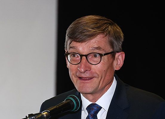 Ein Mann in formaler Kleidung spricht in ein Mikrofon. Er trägt eine Brille und eine Krawatte, vor ihm steht ein Mikrofonständer.