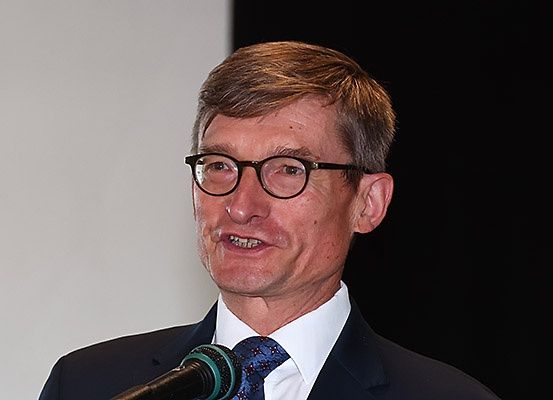 Ein Mann in Anzug und Krawatte, der eine Brille trägt, lächelt und scheint in ein Mikrofon zu sprechen.