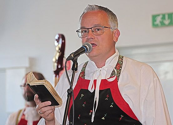 Ein Mann mit Brille und roter Schürze spricht in ein Mikrofon, während er ein offenes Buch in der Hand hält. Ein Mensch mit einer Gitarre steht hinter ihm.