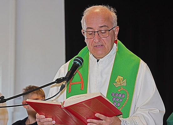 Ein Priester in einer grünen und weißen Stola liest aus einem roten Buch und steht an einem Mikrofon. Ein Junge schaut zu.