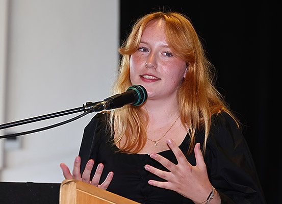 Eine junge Frau mit blonden Haaren steht an einem Rednerpult und spricht in ein Mikrofon. Sie trägt ein schwarzes Shirt und eine Halskette mit Anhänger. Ihre Hände gestikulieren, während sie ein Publikum anspricht.