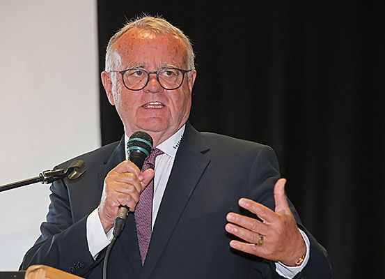Ein älterer Mann in Anzug und Brille steht an einem Rednerpult, hält ein Mikrofon in der Hand und scheint eine Rede zu halten.