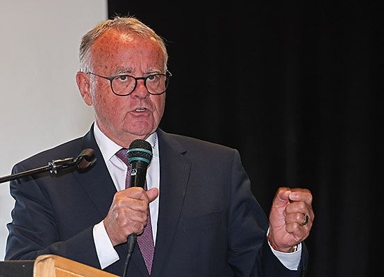 Ein älterer Mann in einem dunklen Anzug, weißem Hemd und lila Krawatte spricht in ein Mikrofon an einem Podium.