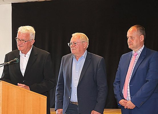 Drei Männer in Anzügen stehen hinter einem Rednerpult. Der Mann auf der linken Seite trägt eine Brille und hat die Hände gefaltet. Der Mann in der Mitte trägt ein blaues Hemd und der Mann auf der rechten Seite trägt eine pinke Krawatte.