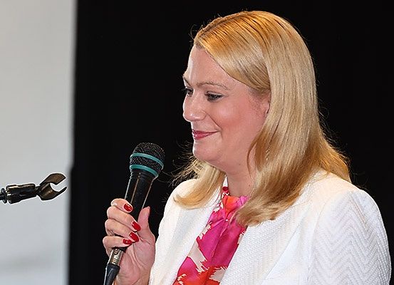 Eine Frau mit blonden Haaren hält ein Mikrofon und scheint zu sprechen. Sie trägt einen weißen Blazer und einen rosa Schal.