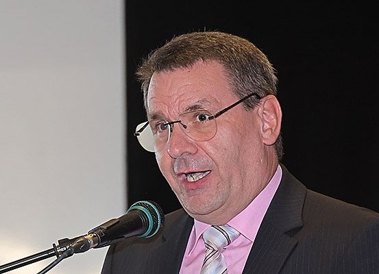 Ein Mann mit Brille spricht in ein Mikrofon, gekleidet in Anzug und Krawatte.