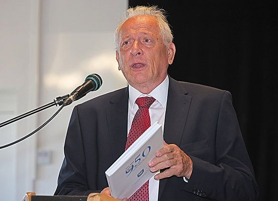 Ein älterer Mann steht an einem Rednerpult, spricht in ein Mikrofon und hält ein Buch mit dem Titel '950' in der Hand.