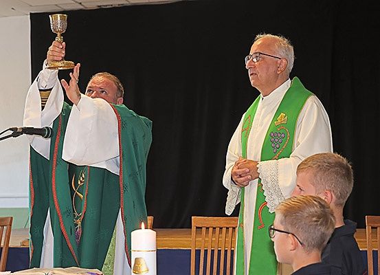 Zwei Priester in grünen Gewändern, einer mit einem Kelch, stehen vor einem Altar mit einer Kerze. Ein Mikrofon ist in der Nähe. Mehrere Kinder beobachten.