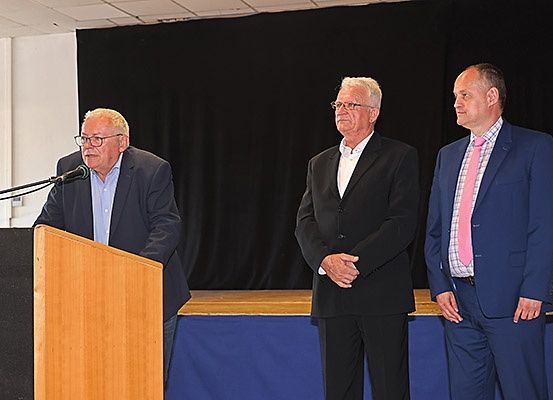 Drei Männer in Anzügen stehen auf einer Bühne. Einer spricht an einem Podium mit einem Mikrofon. Ein schwarzer Vorhang und eine Holzplattform sind im Hintergrund.